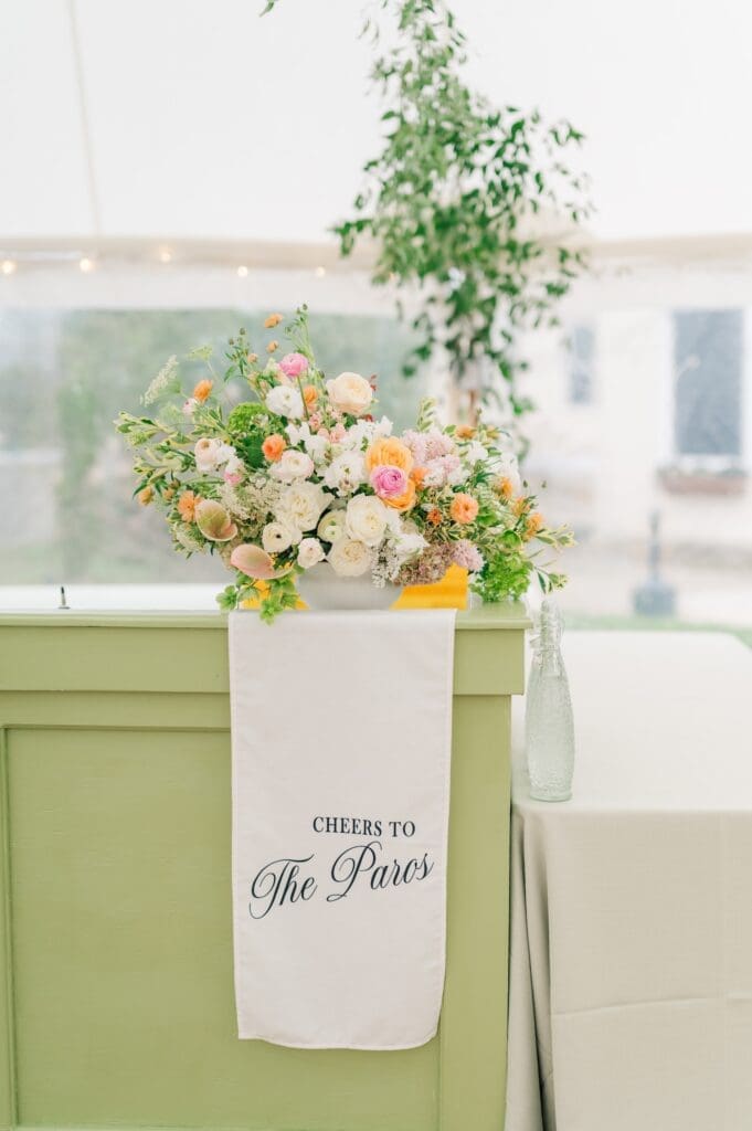 A linen hanging bar sign that says "cheers to The Paros" with a floral arrangement holding it. 