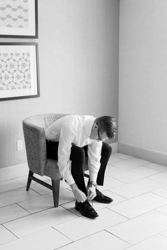 A groom sits and leans over as he ties his shoes. 