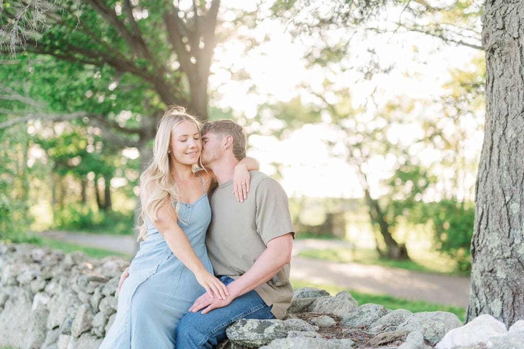 A woman sits on her fiance's lap as he nuzzles into her neck and she smiles. 