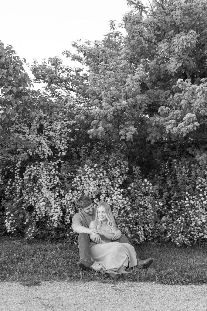 Lois and George sit on the grass in front of a bloomed floral bush and nuzzle into one another. 