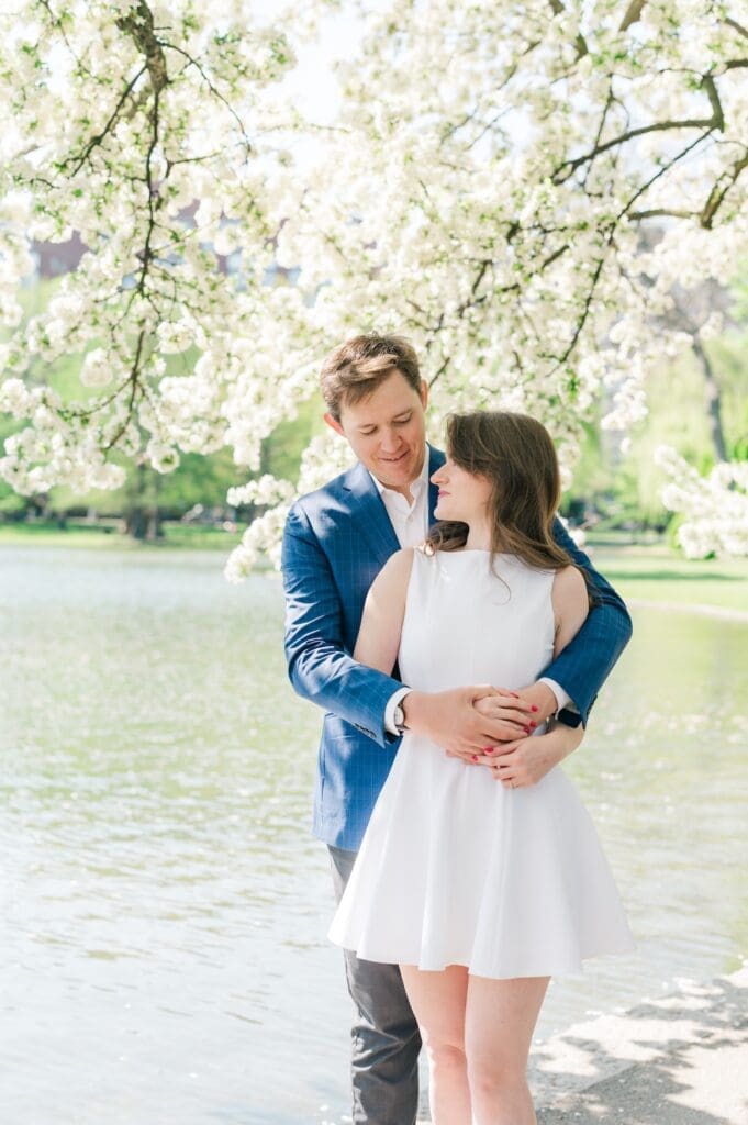 Brandon hugging Julia from behind as they stand under a white blossoming tree in the Boston Public Garden for their engagement photos. 