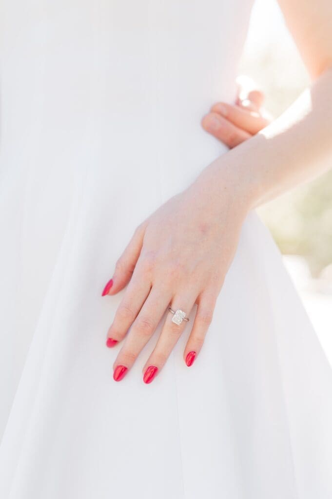 A hand with nails painted red and a radiant engagement ring on the ring finger 
