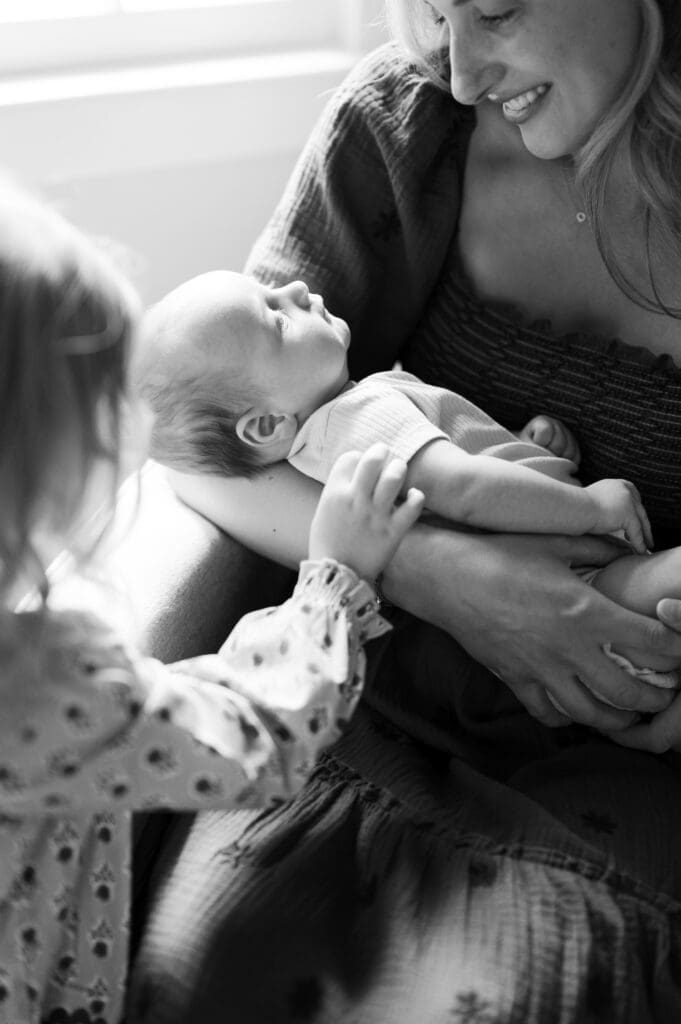 Little sister goes up to mom as she holds baby G and touches his arm