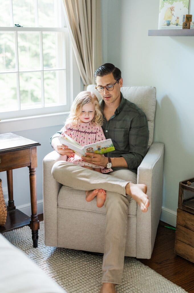 Dad reads with their toddler daughter on their chair. 