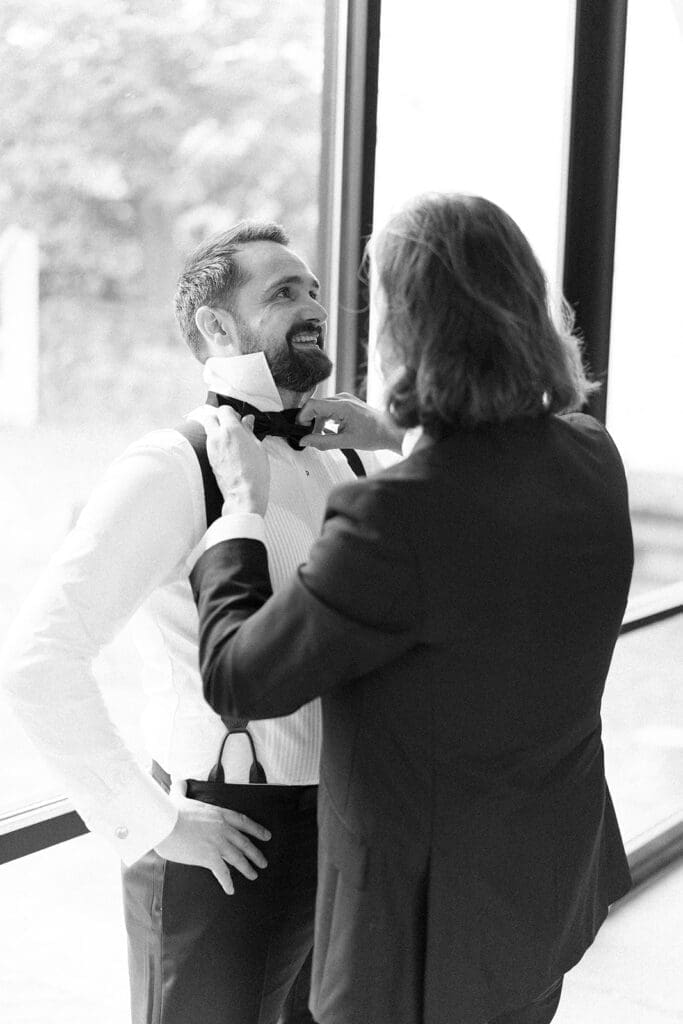A groomsman ties Tom's bowtie indoors on his rainy wedding day 