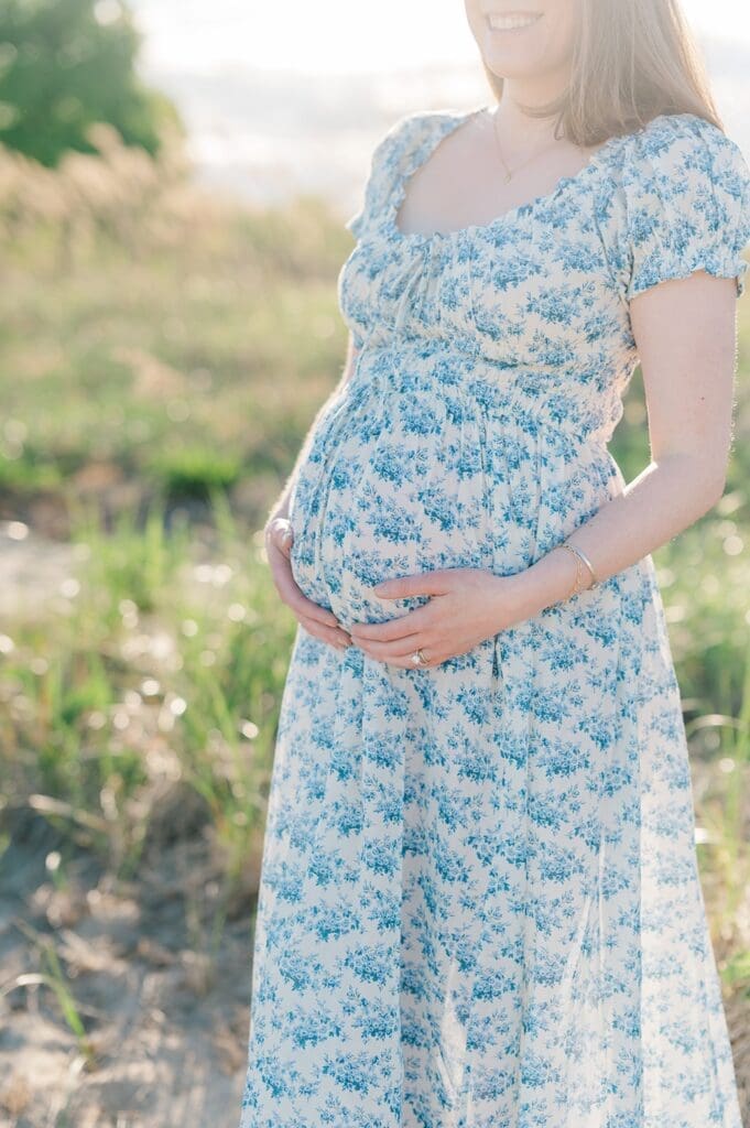 A mother holding her pregnant belly as the sun peeks out from behind her. 