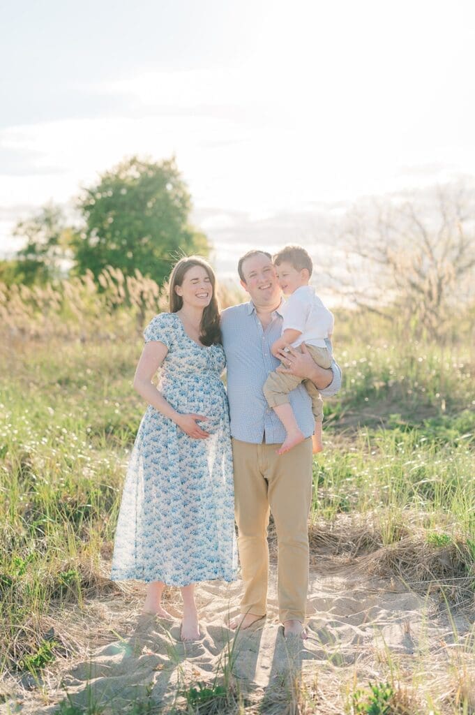A man and woman stand in tall grass on the beach and smile while they hold their toddler son. 