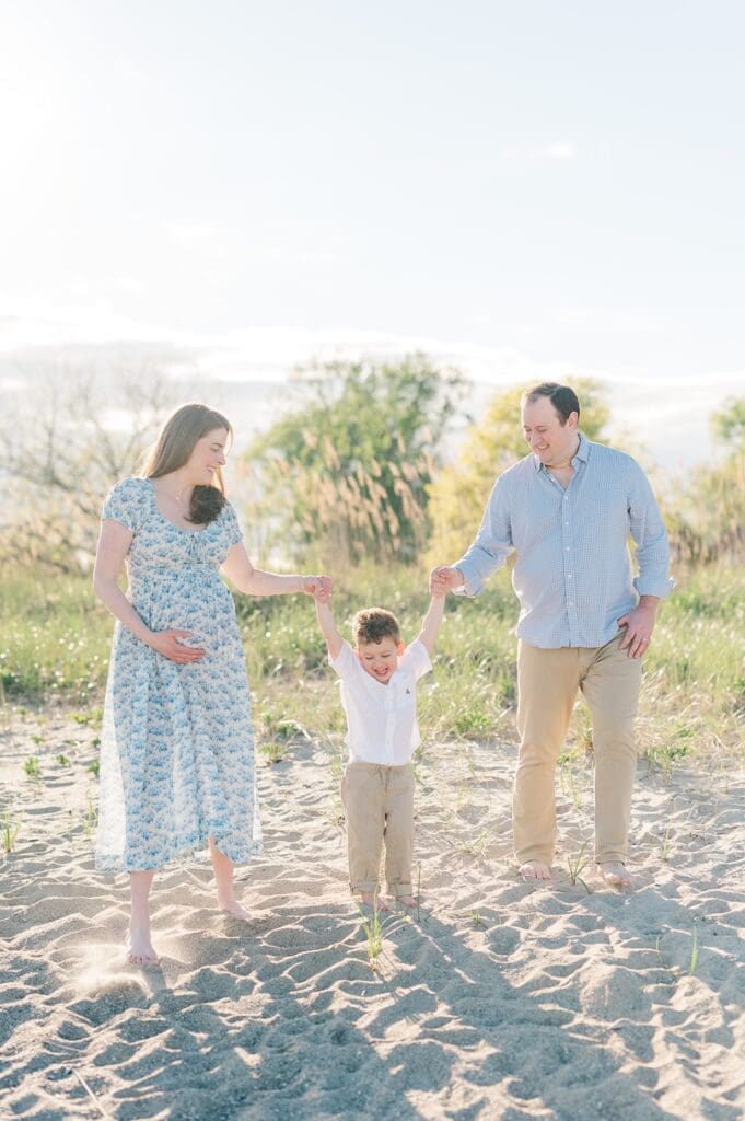 A mom and dad hold hands with their toddler son and walk on the beach laughing together. 