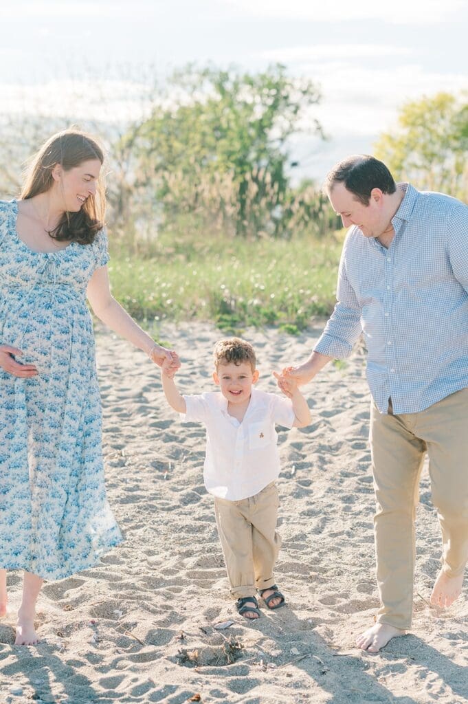 A toddler smiles as his parents hold his hands. 