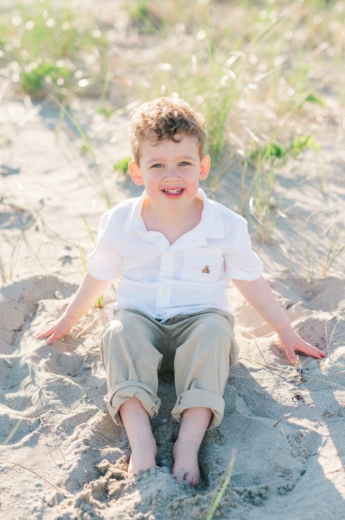 A young boy sits on the sand and smiles candidly. 