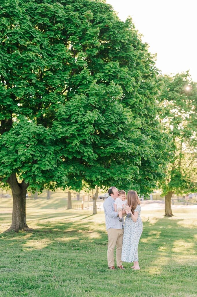 Two parents kiss their child as he laughs. They're standing under a tree in Sherwood Island State Park. 