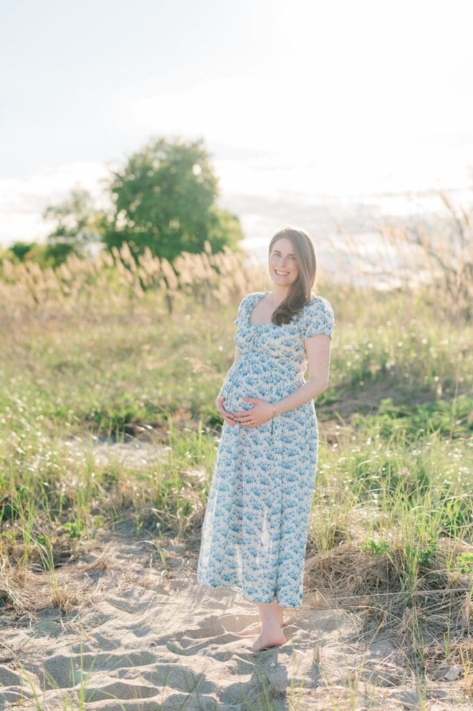 A woman holds her pregnant belly and smiles as the sun sets behind her. 