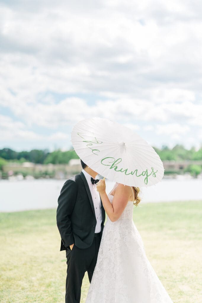 Katie and Sang kiss while they hold a parasol that says "The Chungs".