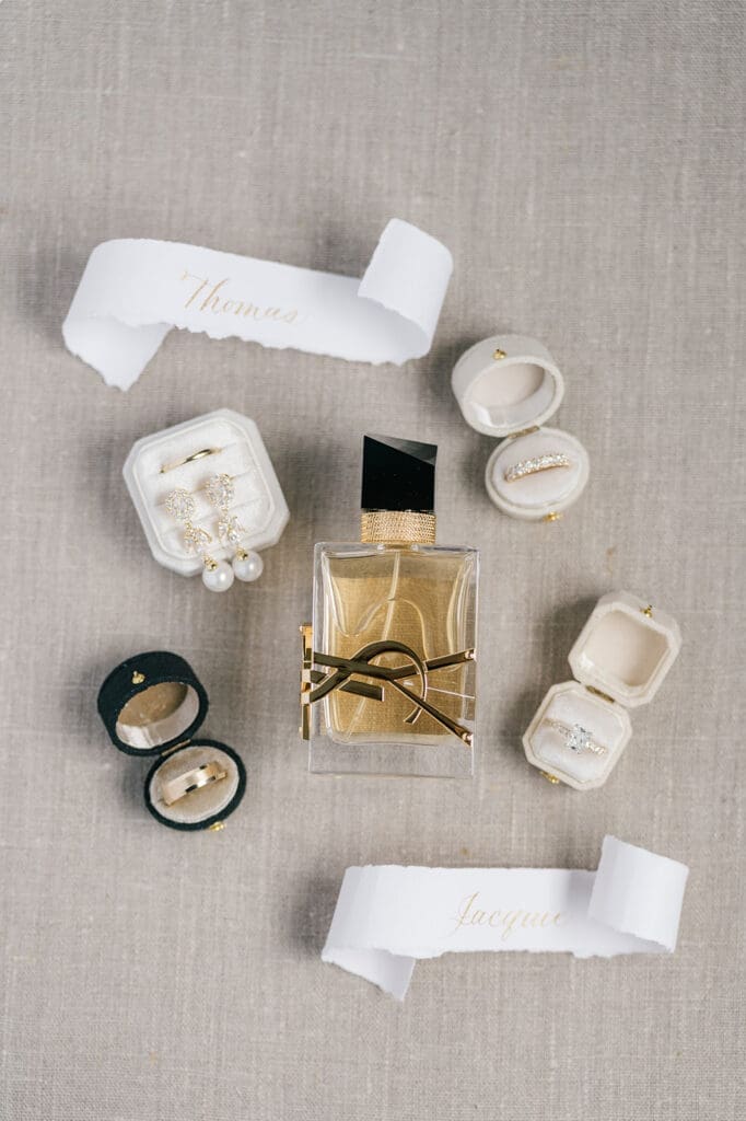 A wedding flat lay of wedding rings, YSL perfume, earrings, and two scrolls that say Jacquie and Thomas. 