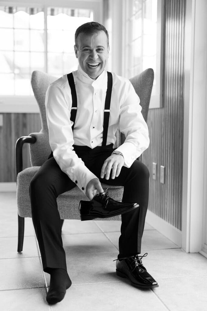The groom, Thomas, puts on his shoes and smiles widely. 