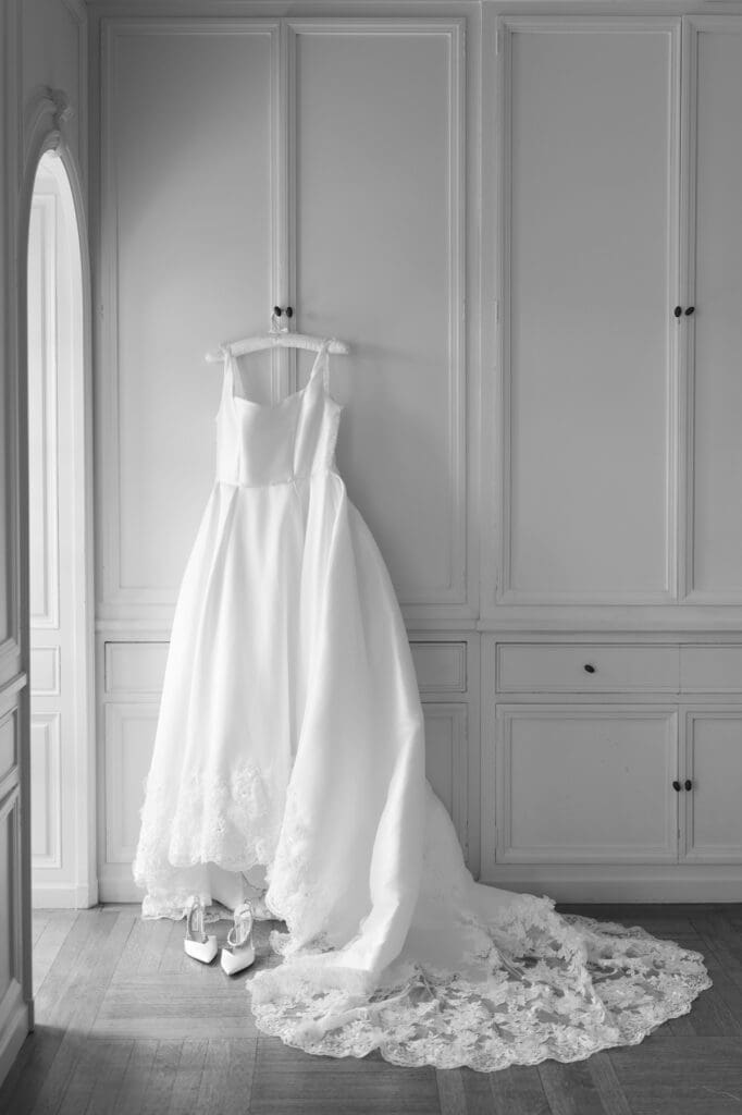 A wedding dress hangs next to a window with shoes below them. 