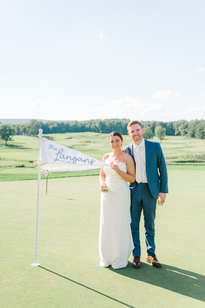 Amy and Conor stand next to a golf flag that says "The Langans" at their Wampanoag Country Club Wedding