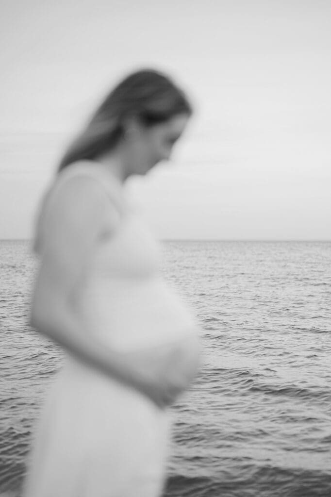 Lindsey holds her belly as the focus is on the ocean water behind her.