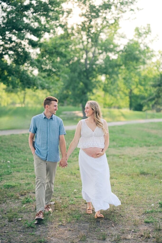 Rich and Lindsey hold hands and walk under trees at Sherwood Island State Park for their maternity photoshoot in CT.