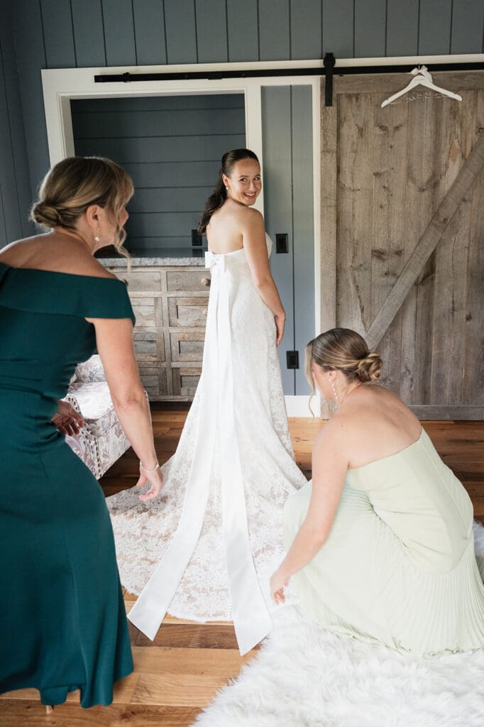A bride looks back at her mom and sister as they adjust her dress. 