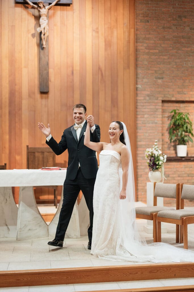 A bride and groom smile and hold their hands in the air as they cheer after getting married in the chapel. 
