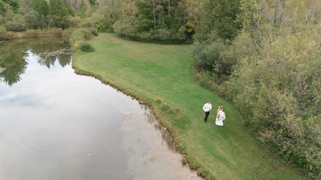 A drone photo of a bride and groom walking in the grass together next to a pond. 