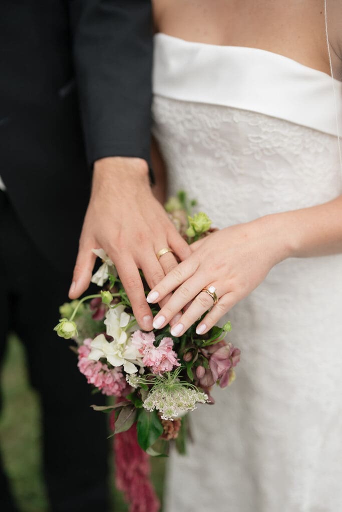 A bride and groom hold their ring fingers over the bride's bouquet. 