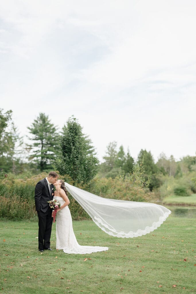 A bride and groom kiss as the bride's veil flies in the wind behind them at their Barnstead Inn wedding. 