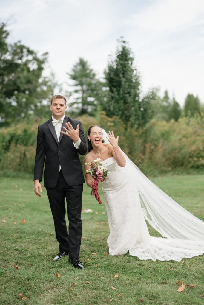A bride and groom hold up their ring hands and smile. 