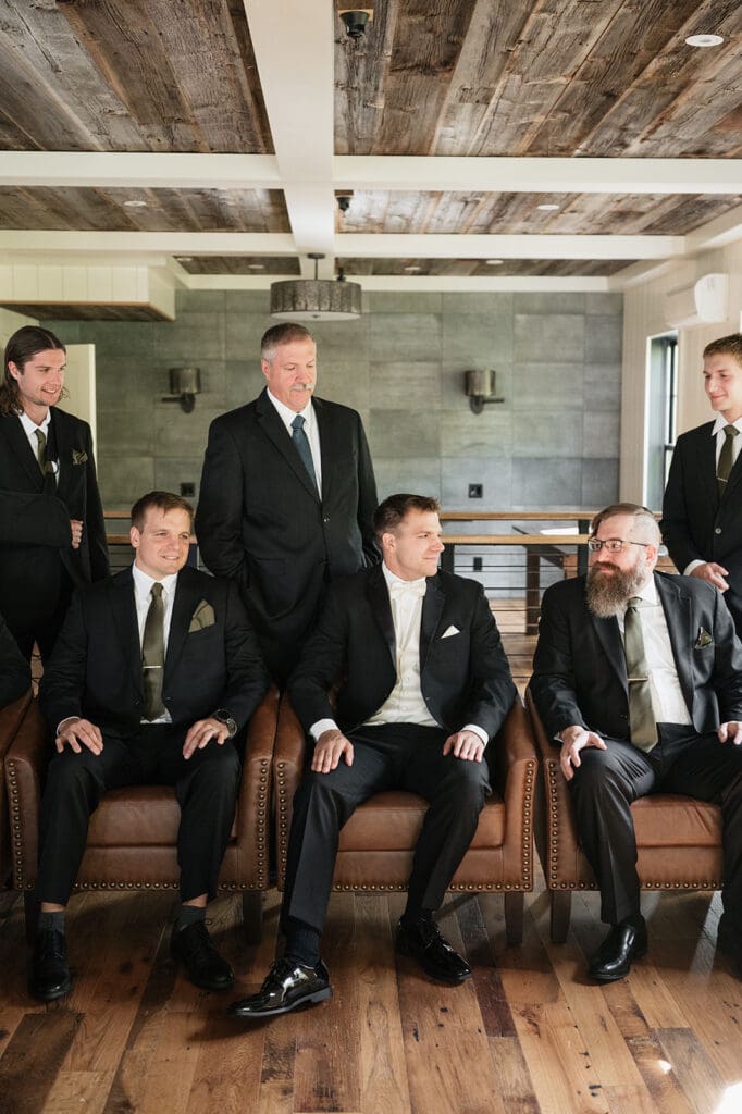The groom and his groomsmen sit on chairs and smile at eachother. 