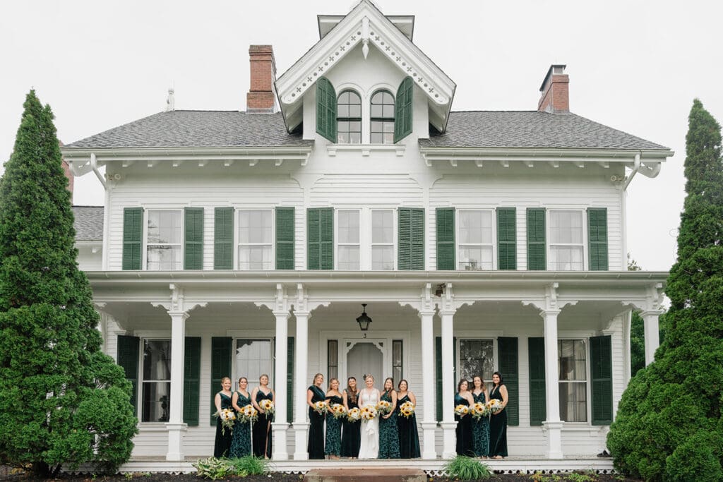 A bride and a group of bridesmaids stand on the front porch of the Lyman Orchards Homestead for a group photo
