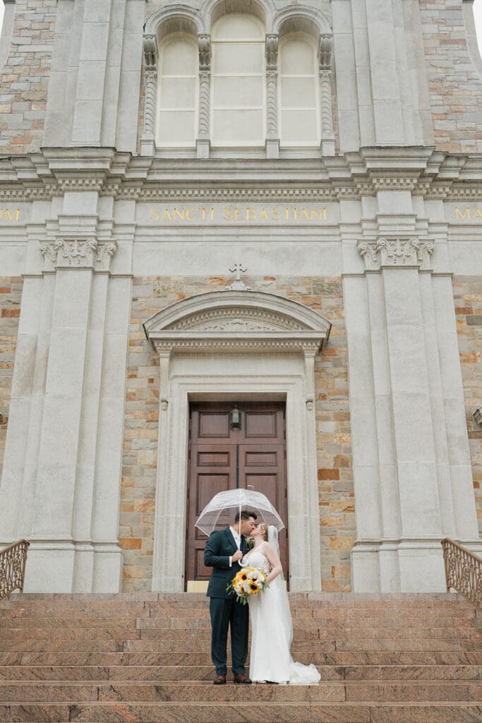 A bride and groom kiss under an umbrella as it rains on their wedding day.