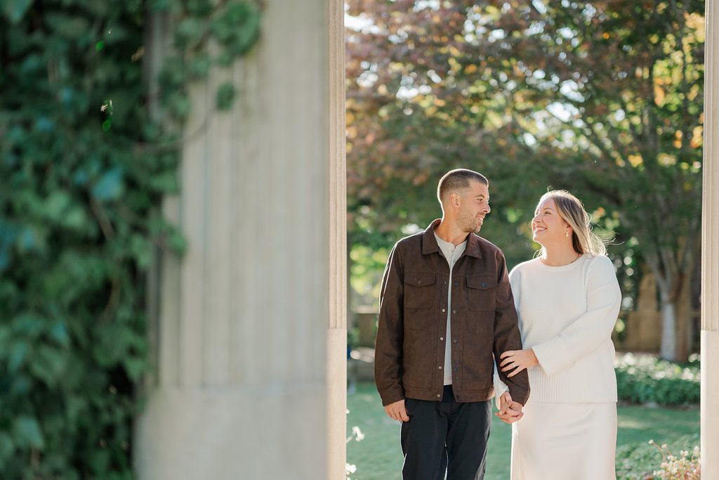 Engaged couple walking hand in hand through the stone pergola columns at Harkness Memorial State Park, with fall trees behind them