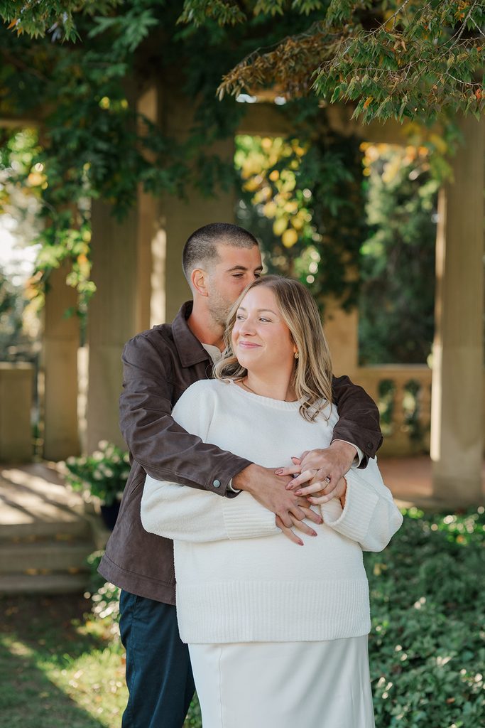 Fiancé hugging his fiancée from behind and leaning in close under the pergola, with fall leaves overhead, at Harkness Memorial State Park, CT