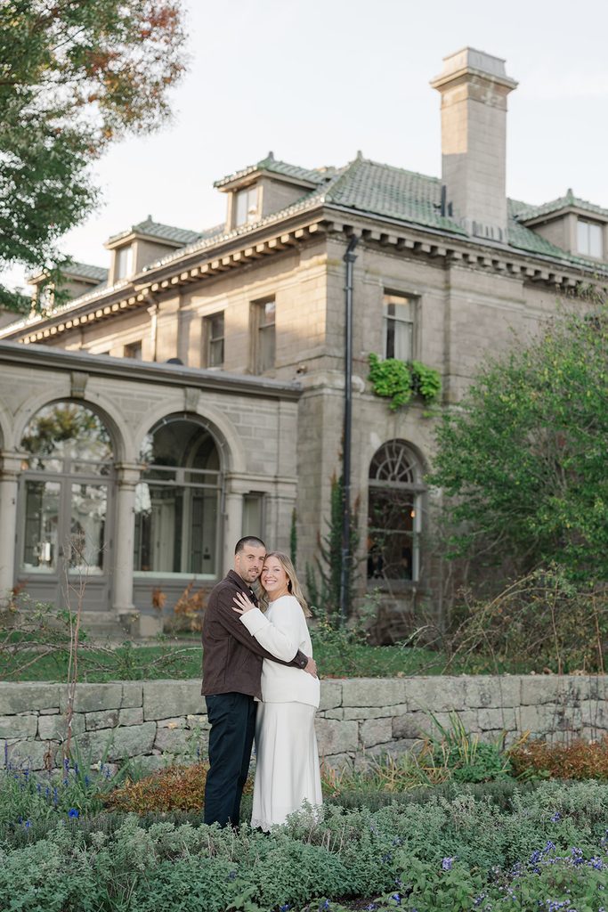 Engaged couple embracing in the gardens in front of the Eolia mansion at Harkness Memorial State Park in Waterford, CT