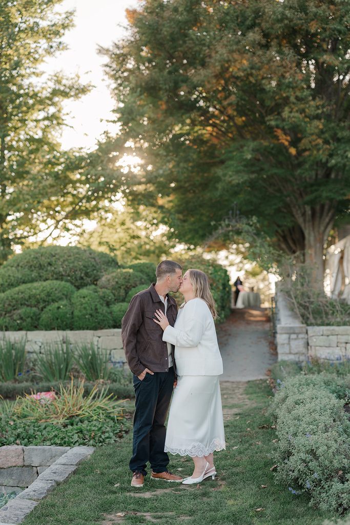 Engaged couple kissing on a grassy garden path between boxwood hedges and stone walls at Harkness Memorial State Park, CT