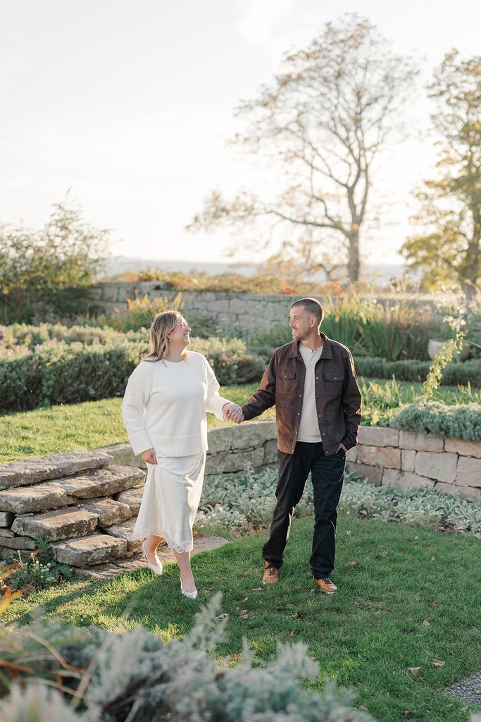 Engaged couple holding hands and smiling at each other on the garden lawn by a stone wall, with Long Island Sound behind them at Harkness Memorial State Park