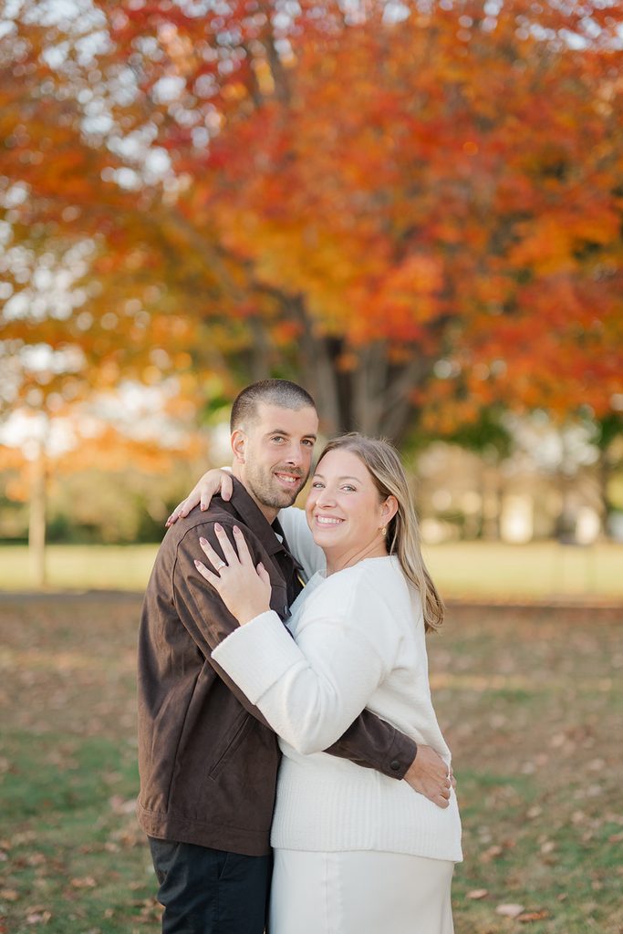 Engaged couple hugging and smiling at the camera in front of a bright red and orange fall maple at Harkness Memorial State Park