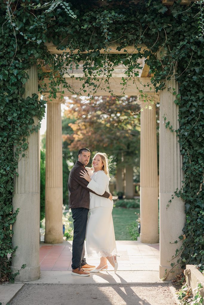 Engaged couple embracing under an ivy-covered stone pergola at Harkness Memorial State Park in Waterford, CT, with fall foliage behind them