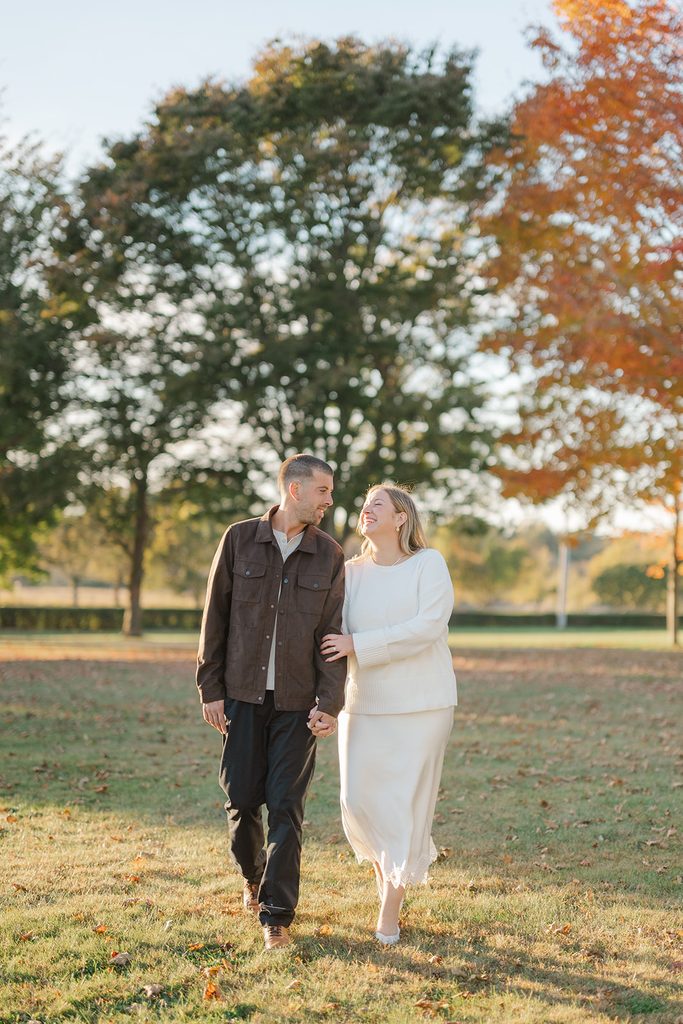 Engaged couple walking hand in hand across the lawn at golden hour with orange fall trees during their engagement session at Harkness Memorial State Park, CT