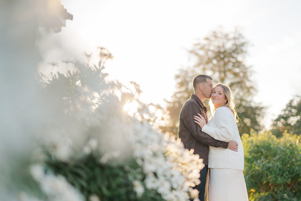 Fiancé kissing his fiancée's temple at golden hour beside white flowers during a fall engagement session at Harkness Memorial State Park, Connecticut