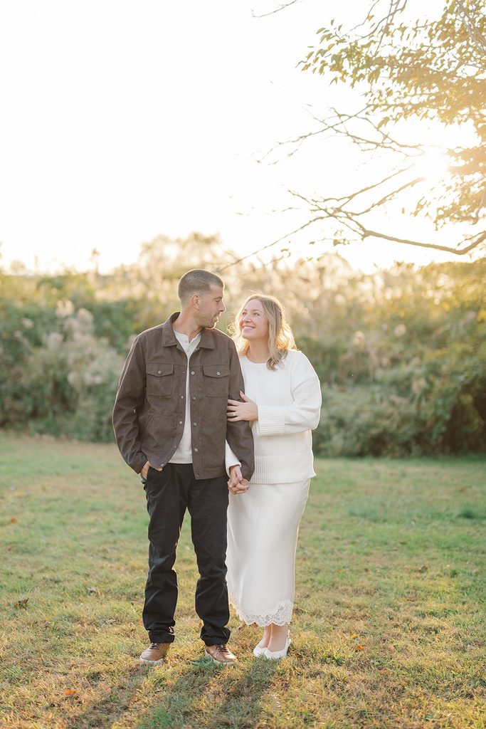 Engaged couple holding hands in a sunlit meadow at golden hour, she smiling up at him, at Harkness Memorial State Park in Waterford, CT