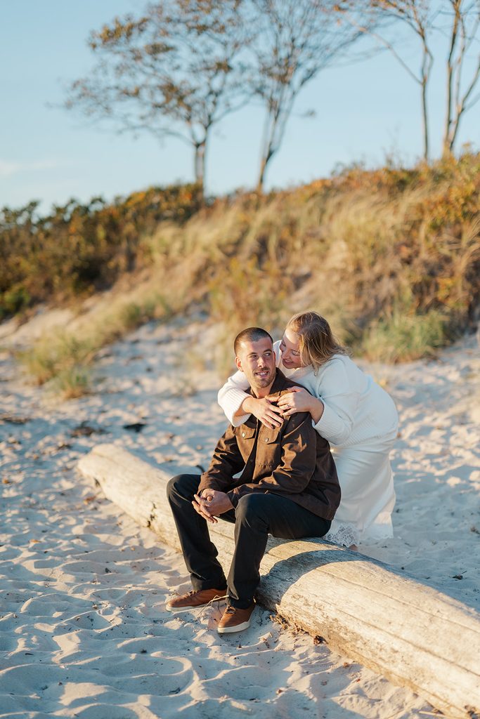 Engaged couple on a driftwood log on the beach at Harkness Memorial State Park, she hugging him from behind, with dune grasses in fall color