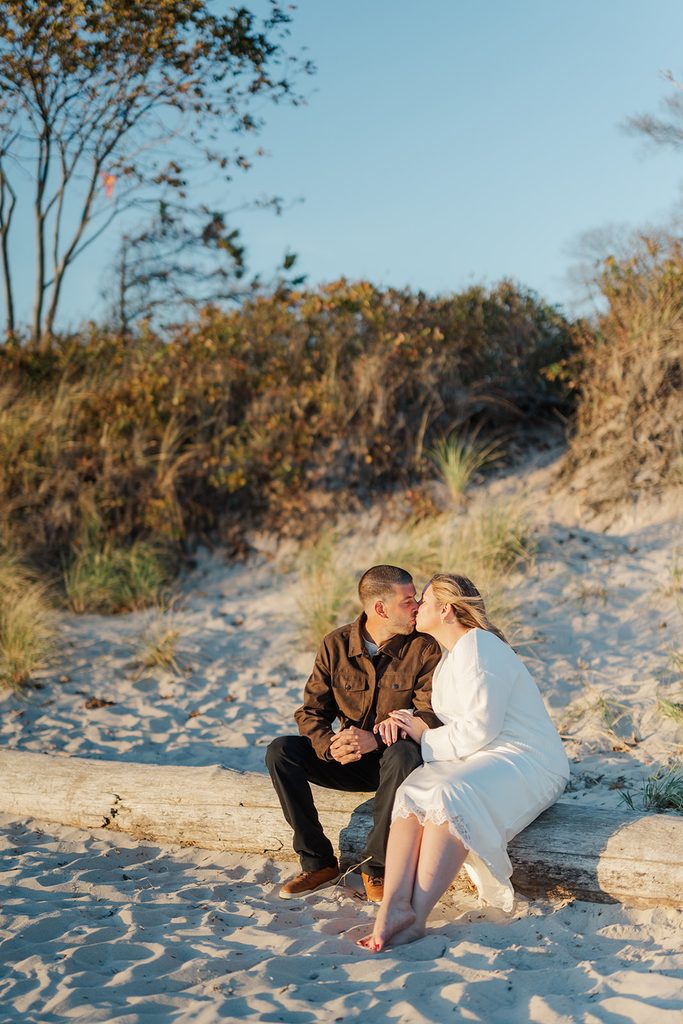 Engaged couple kissing on a driftwood log on the sandy beach at Harkness Memorial State Park, with fall dune brush behind them