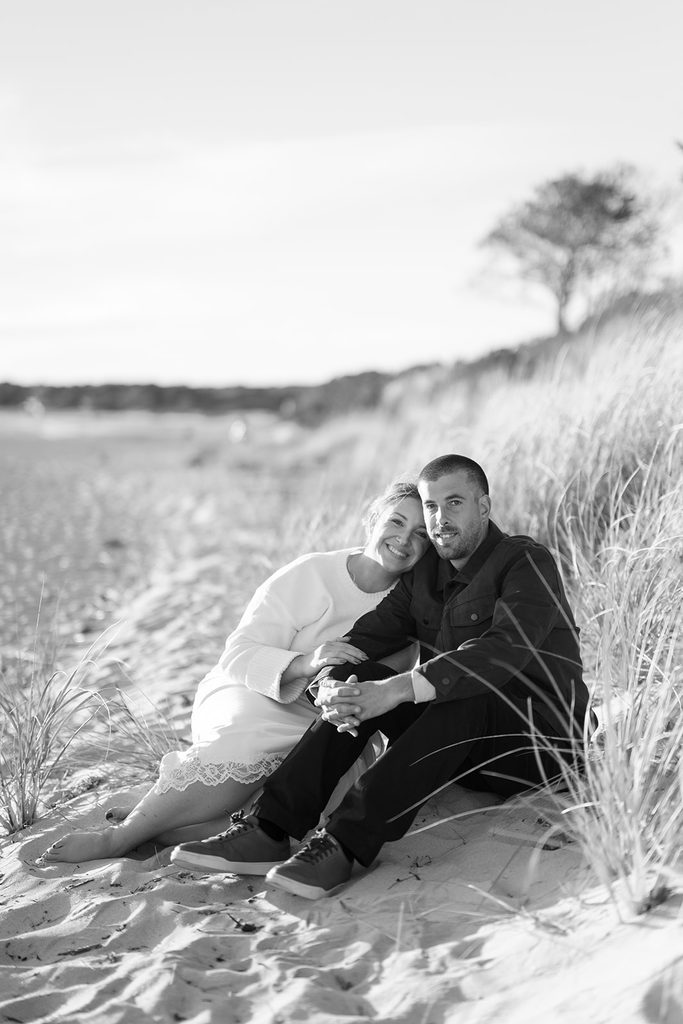 Black and white photo of an engaged couple sitting together in the beach grass and smiling at the camera at Harkness Memorial State Park