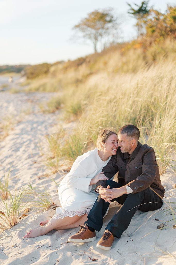 Engaged couple sitting in the beach grass on the dunes at Harkness Memorial State Park, laughing with their foreheads touching at golden hour