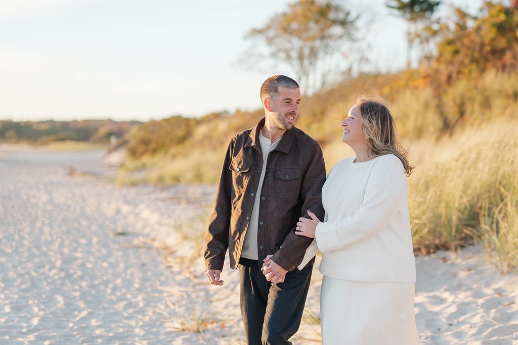 Engaged couple walking arm in arm along the beach at golden hour, smiling at each other, at Harkness Memorial State Park, Connecticut