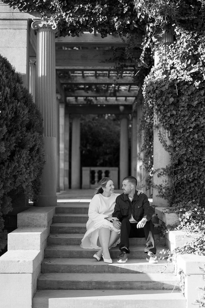 Black and white photo of an engaged couple sitting on stone steps beneath the pergola at Harkness Memorial State Park, smiling at each other