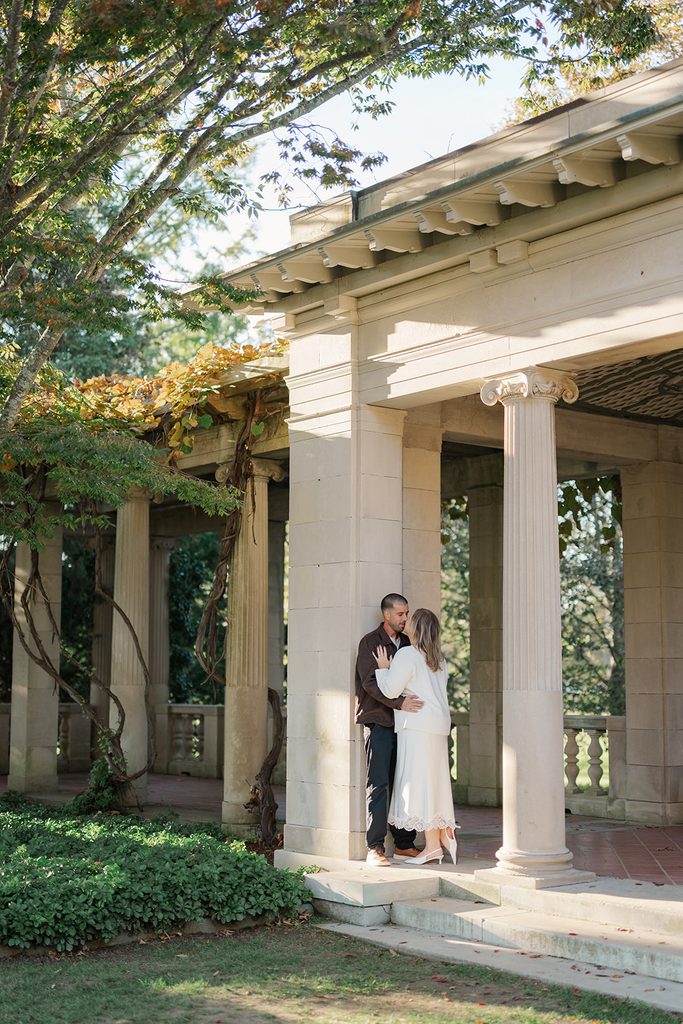 Engaged couple kissing against a stone column of the pergola at Harkness Memorial State Park, with vines and dappled sunlight